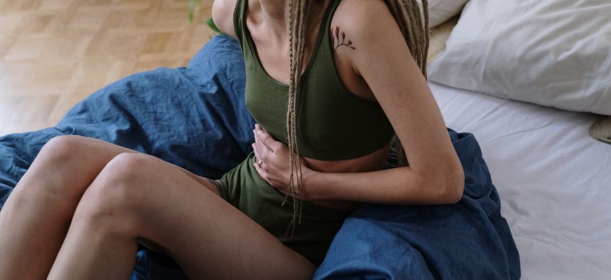 Woman sitting in bed holding her stomach, indicating pain or discomfort, possibly due to cramps.